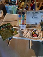 Art supply set on a small white folding table showing wooden MÅLA easel, multiple watercolor paint sets including rectangular palette and green-case compact set, and white plastic paint palette.