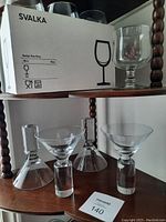 Photo showing a box labeled SVALKA with 6 wine glasses and 4 martini glasses displayed on wooden shelving.