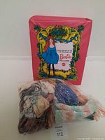 Closed pink carrying case with vintage Barbie illustration and a large bag of assorted Barbie doll clothes in plastic bags