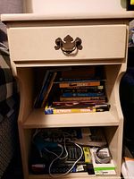 Front view of the antique white Ethan Allen side table showing one drawer filled with several books, and two lower shelves containing various household items and electronics cords. Drawer has ornate metal handle.