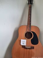 Front view of the acoustic guitar showing 6 strings, natural wood body, black pickguard, and bridge with white pins.