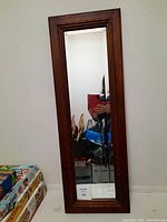 Photo showing the mirror standing vertically against a white wall, reflecting nearby objects including some boxed items and a person taking the photo.