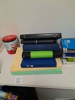 Stack of six binders, large sheets of green and yellow paper, and a red round tin on a white table against a plain wall.