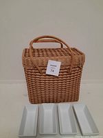 Photo showing a woven rectangular picnic basket with a curved handle and four white rectangular bowls placed beside it, against a plain background.