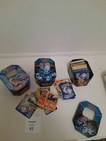 Three Pokemon card canisters with assorted cards visible inside and some loose cards outside each tin.