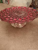 Side angled photo of red glass cake plate showing floral petal design, pedestal base, and textured finish.