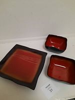 Three-piece set of ceramic tableware with one large square serving plate and two smaller square bowls, red with black edges and subtle design on the plate.
