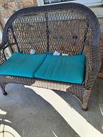 Image showing brown wicker two seater patio sofa with teal green cushions on concrete floor in front of house wall.