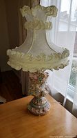Front view of the ornate porcelain table lamp showing detailed Baroque style floral and scroll design on the base with fabric shade featuring ruffled edges.