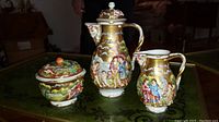 Hand-painted porcelain coffee pot, creamer, sugar bowl shown on table, decorated with gold and classical scenes