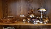 Wide view of all pieces showing variety of depression glass items with metal accents including tall covered serving dish, decanter, small bowls, and cordial glasses.