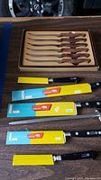 Six steak knives with wooden handles in a box, displayed above five Sabatier cutting knives in original packaging.