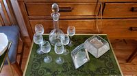 Etched glass decanter with stopper, six dessert wine glasses, glass candle holder with candle on green tray in front of wooden dresser.