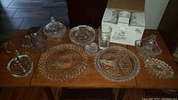 Image showing the assortment of clear pressed glassware including serving platters, candy dishes with lids, a pitcher, and box of six tall glasses.