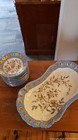 12 small plates stacked beside a large irregularly shaped bowl/platter, both decorated with blue and gold floral rims and brown floral patterns.
