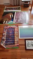 Photo showing five posters laid out on a wooden floor including Summerfair 2004, wildlife walrus poster, purple portrait, and a framed black and white art piece with geometric Native American motifs.