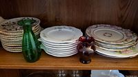 Photo of shelves showing stacks of various china plates and two small decorative glass vases, one green and one red.