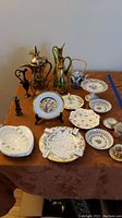 Wide view of lot showing assorted vintage collectibles on brown tablecloth including glass pitchers, porcelain plates, ashtrays, teapot, and candlesticks