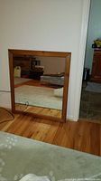 Front view of the wood frame mirror resting against a wall on a hardwood floor, showing the frame and reflective glass.