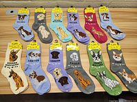 Top-down photo showing all 12 new dog socks laid out flat on wooden surface with attached packaging tags visible. Each sock features a colorful cartoon dog breed illustration and breed name.