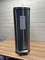 Front view of tall Brightroom stainless steel trash can on wooden table against brick wall.