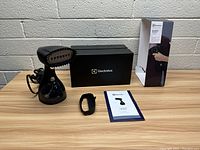 Full view of the Electrolux handheld steamer with its brush attachment, user manual, fabric strap, and both the product box and branded black box on a wooden table