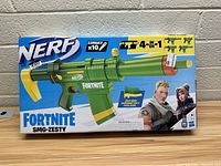Front view of Nerf Fortnite SMG-Zesty toy blaster box on wood surface against a concrete wall showing product image and Fortnite characters.