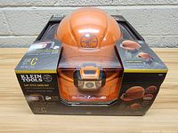Front view of the orange Klein Tools cap-style hard hat in original packaging showing the product and headlamp.