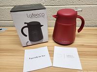 Photo showing red Lafeeca stainless steel carafe next to its original box and two informational cards on a wooden surface.