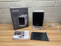 Levoit Fresh Air HEPA Air Purifier with original box, sealed replacement filter pad, and instruction manual laid out on a wood surface against a light brick wall.