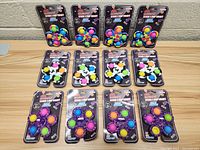 Photo showing 12 packaged Stick & Pop Fidget toys arranged standing against wall on wooden table, displaying front packaging and colorful pop bubbles visible.