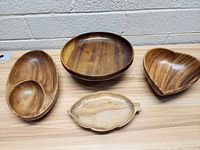 Four wooden bowls of various shapes including heart, oval with compartments, round, elongated oval, and one small beige ceramic leaf-shaped dish with handle, all displayed on a wooden surface.