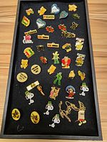Top view of assortment of collectible enamel pins spread on black fabric, showing various designs including Disney characters, Gumby, Coca Cola, Elvis, and Snoopy pins.