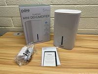 Photo showing the Pure Enrichment PureDry Mini Dehumidifier alongside its box, power cord, and instruction manual on a wooden surface.