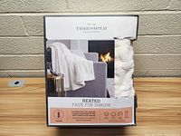 Front view of the Threshold heated faux fur throw blanket box showing white plush throw displayed on couch beside fireplace.