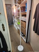 Full view of the white floor lamp standing upright with cylindrical clear glass shade covering a retro filament bulb, set in a narrow room with shelves and a carpeted floor.