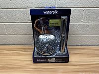 Front view of boxed Waterpik 2-in-1 PowerWash shower system with visible showerhead and handheld wand inside the retail packaging and label details.