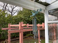 Decorative outdoor metal hanging wind chime shown from a distance hanging under a roof with outdoor garden in background.