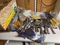 Large assortment of vintage cutlery including spoons, forks and knives with yellow handles on a shelf among boxes and fabric.