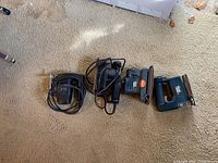 Lot of four tools on carpet including two electric sanders and two electric staplers, cords wrapped around tools.