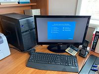 Full setup on wooden desk including black PC tower, Samsung monitor with Windows setup screen, Logitech keyboard, black cordless phone, and white calculator.