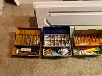Photo showing three fishing tackle boxes filled with assorted fishing lures and accessories. The boxes vary in color and contain multiple compartments with visible lures, fishing line, and packages.