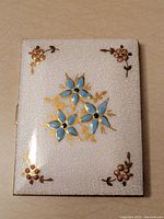 Front exterior of cigarette case showing white crackled enamel surface with blue and gold flower design and corner pink flowers.