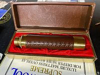 Closed view of vintage brass spyglass wrapped in brown stitched leather, placed inside a brown case lined with red fabric.
