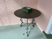 Full view of vintage round metal table with a square green planter on top, showing its aged top surface and wrought iron base with scroll designs.