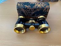 Front view of vintage opera glasses showing black and gold-tone metal body with adjustment knob and accompanying fabric case with gold leaf pattern.