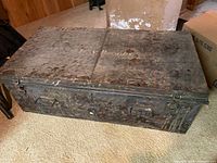 Full view of heavily rusted and worn vintage WWII-era metal trunk placed on carpet floor with side handles visible.