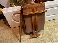 Overall view of folded wooden artist's easel showing hinges, support ledge, and adjustable brass hardware.