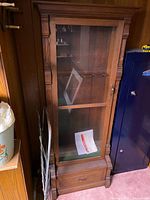 Front view of full-height wooden display cabinet showing glass-paneled hinged door, fluted side columns, decorative top molding and single lower drawer