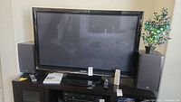 Full front view of the 52-inch Panasonic television on stand with remote control on the stand and two large speakers on each side.